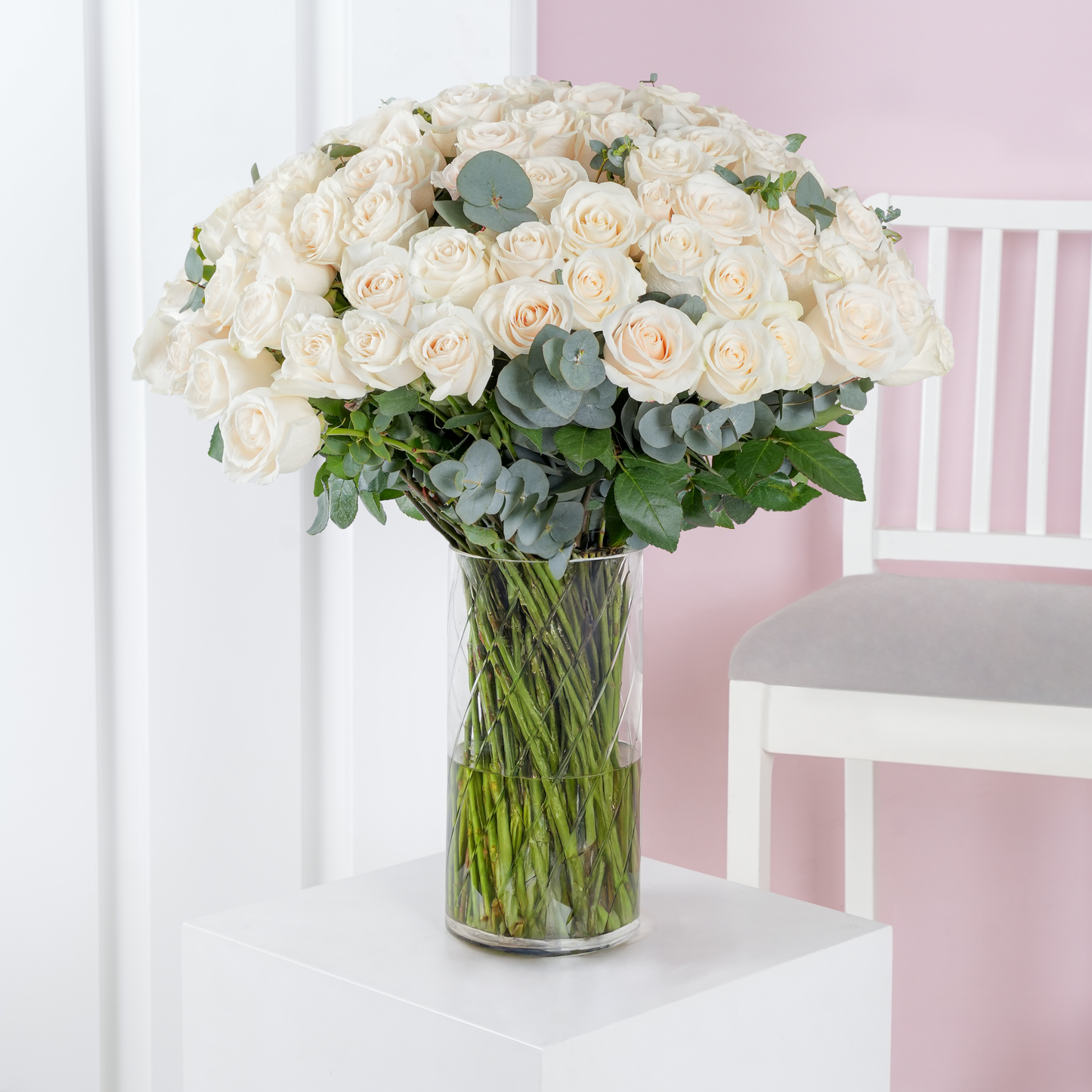100 white roses in vase with green leaves arranged in a clear glass vase on a white pedestal