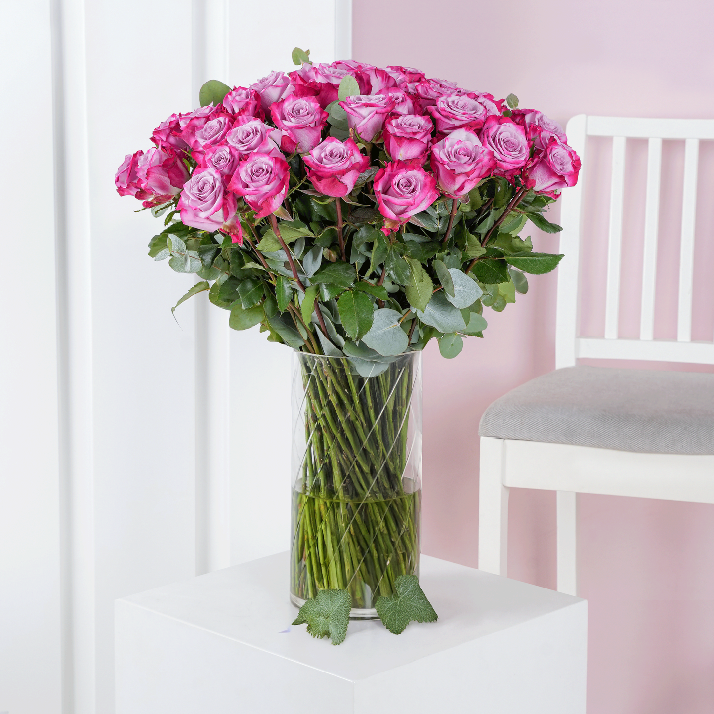 50 purple roses in vase arranged with green leaves on white pedestal next to white chair