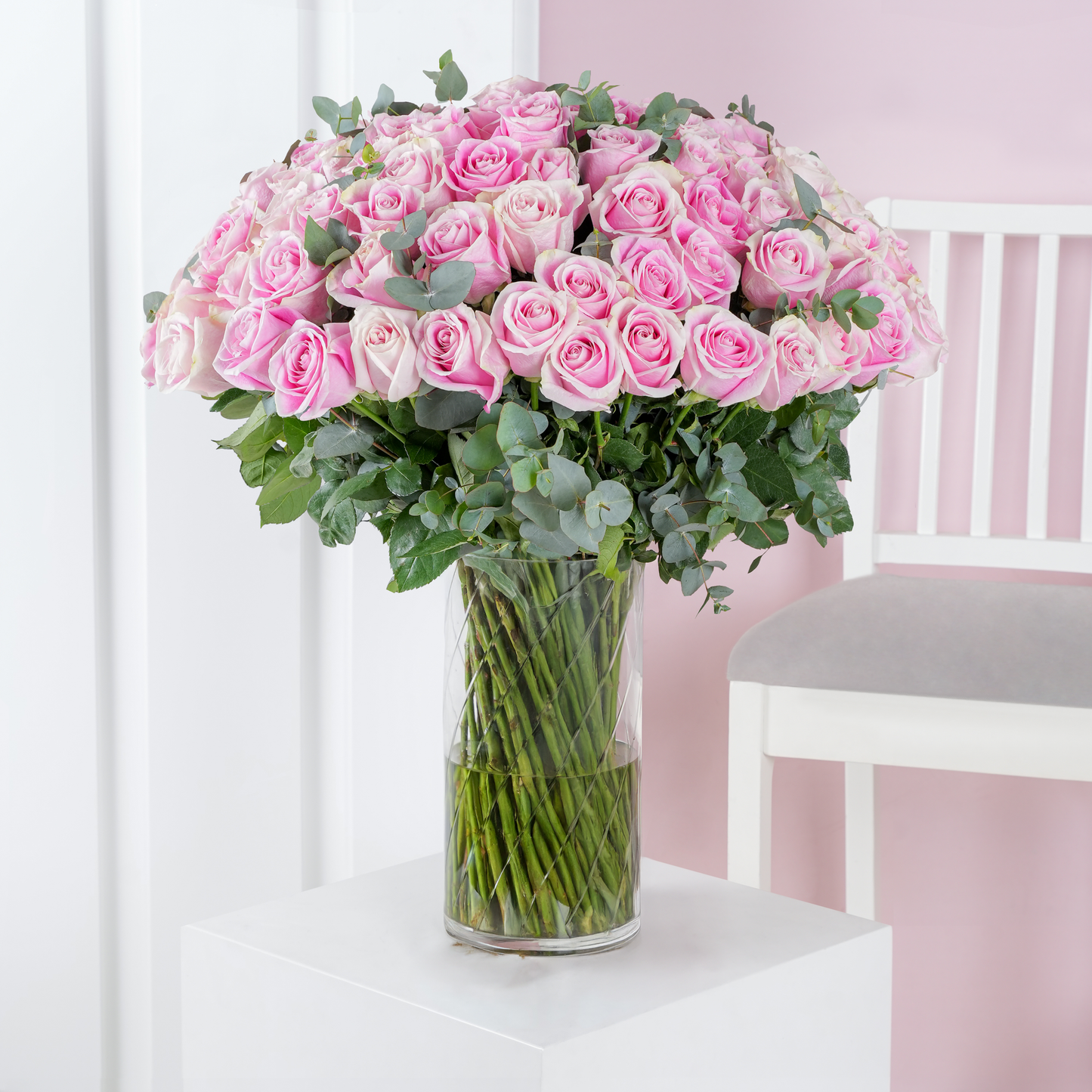 100 pink roses bouquet Bloom Marbella displayed in a clear glass vase with green foliage on a white table