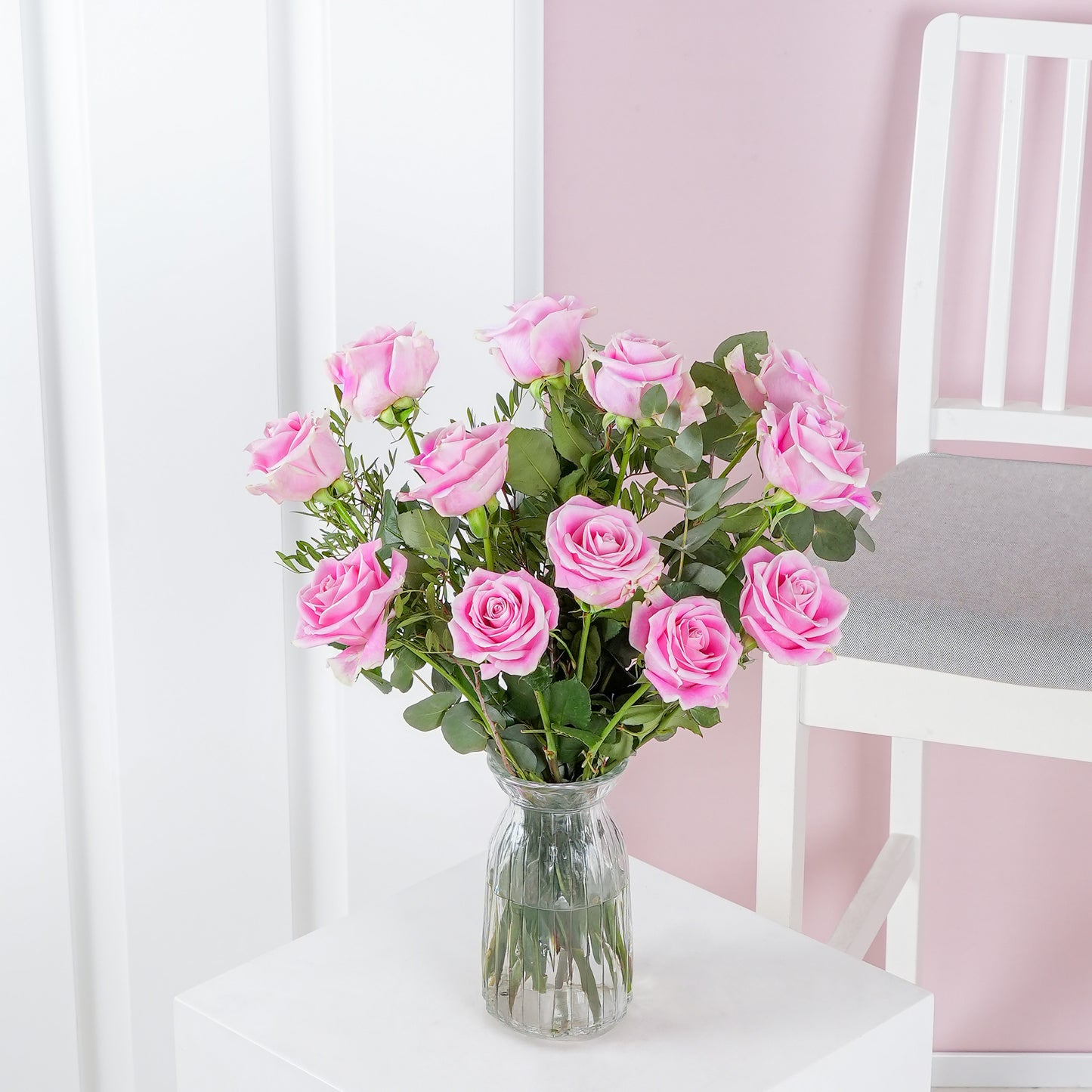 12 pink roses in vase - Bloom Marbella arranged with green leaves in clear glass vase on white table