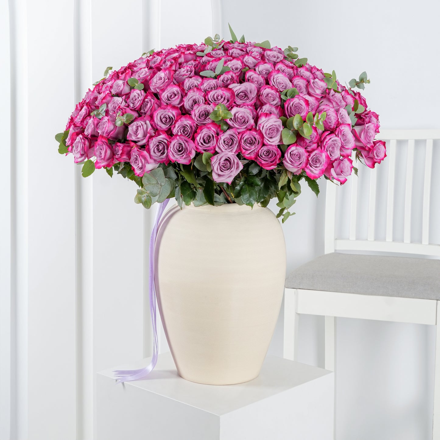100 purple roses in vase - Bloom Marbella, Spain arranged in cream vase with green leaves on white pedestal