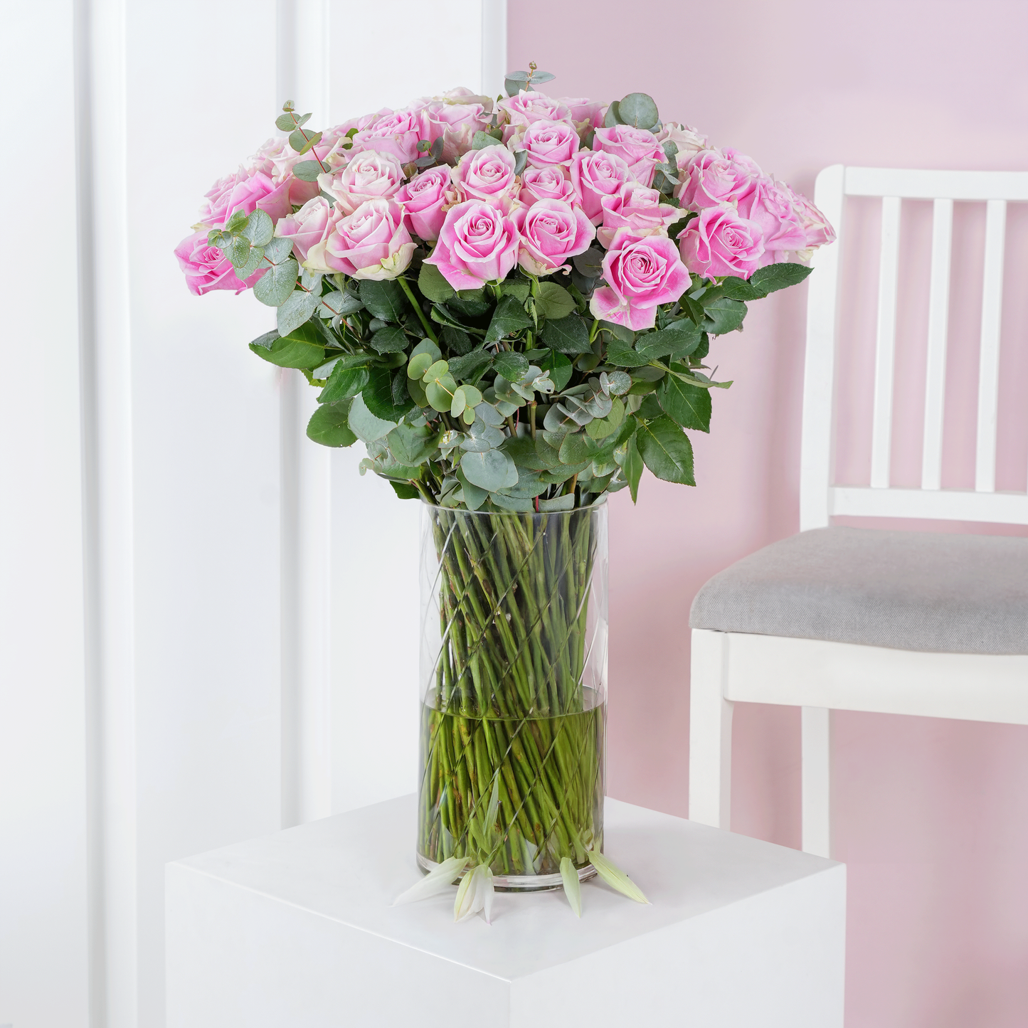 50 pink roses in vase arranged with green leaves in a clear glass vase on white table next to white chair