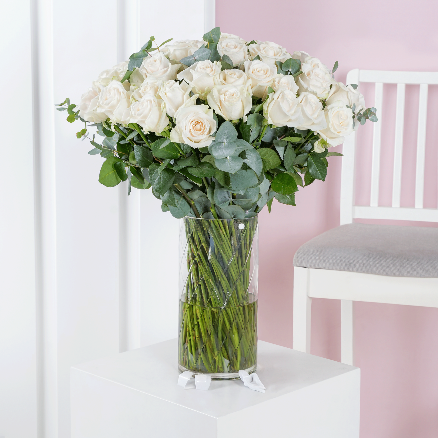 Elegant white roses bouquet Marbella arranged in a clear glass vase with green leaves on white pedestal