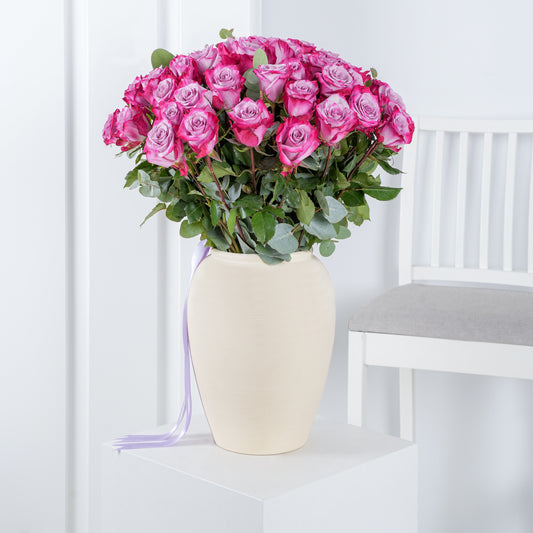50 purple roses in vase arranged with green foliage in a cream ceramic vase on white pedestal