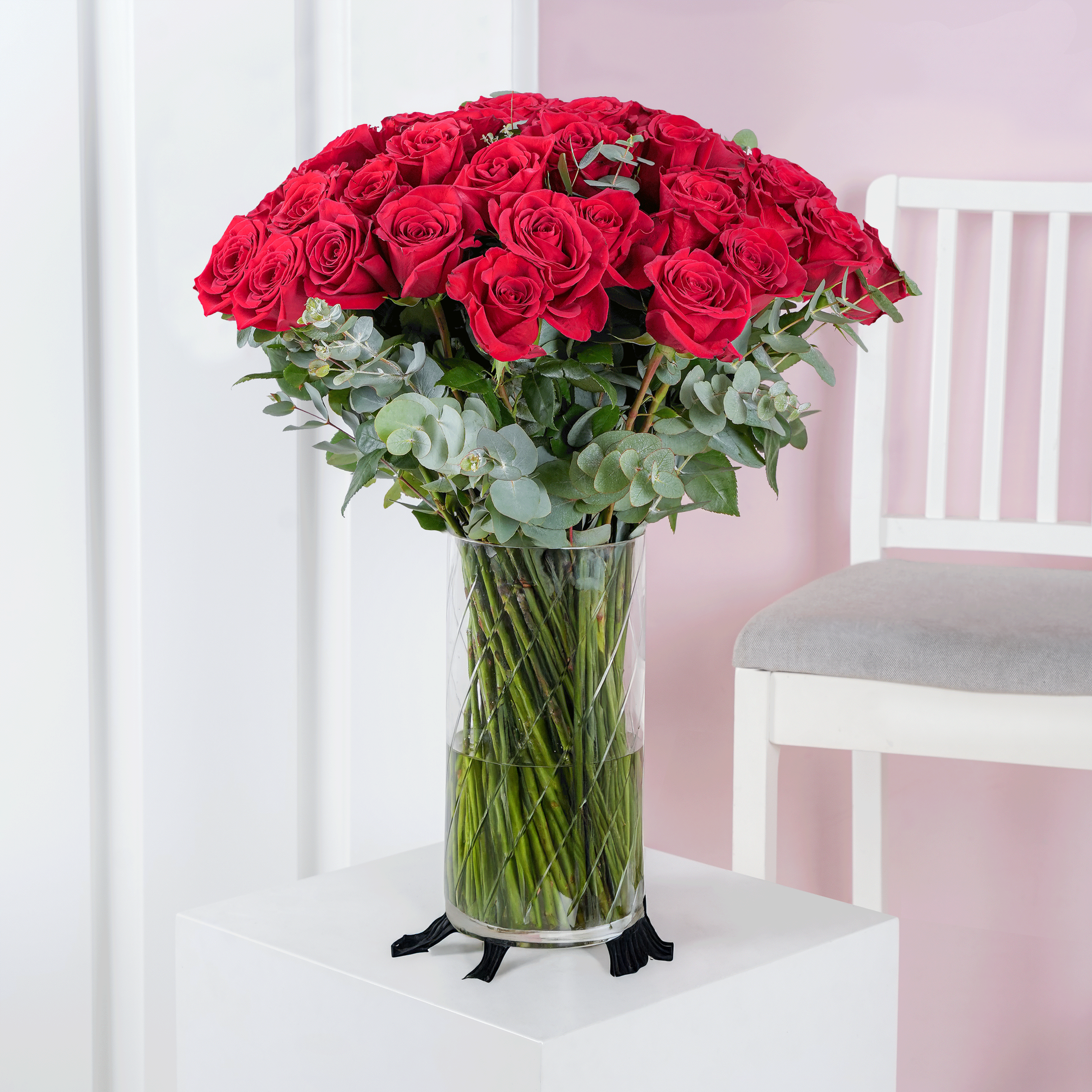 50 red roses in vase arranged with green foliage on white table next to white chair