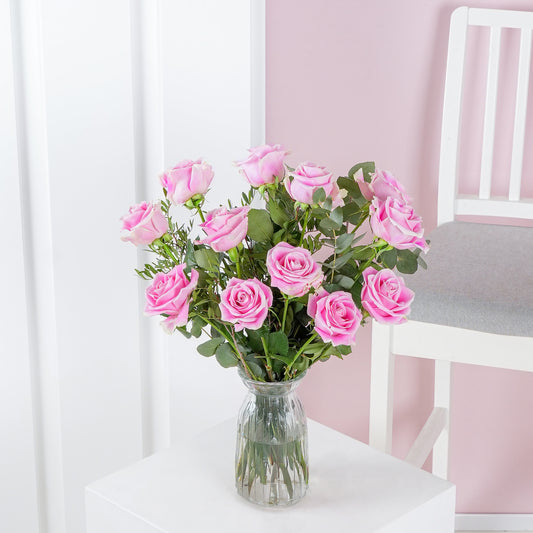 12 pink roses in vase - Bloom Marbella arranged with green leaves in clear glass vase on white table