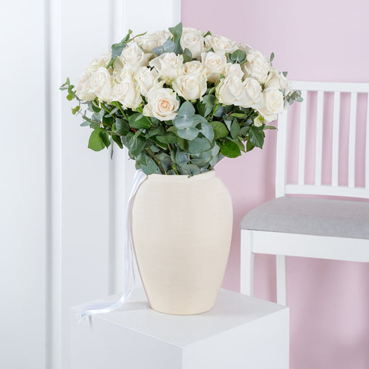 White roses bouquet Marbella arranged with greenery in a cream vase on a white pedestal